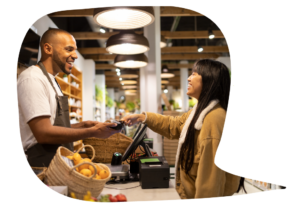 A man behind a counter at a food store smiles as he accepts a payment card from a smiling woman making a purchase