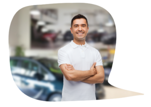 A man in a short sleeved white shirt stands with arms crossed and smiling in a car showroom with a vehicle just behind him.