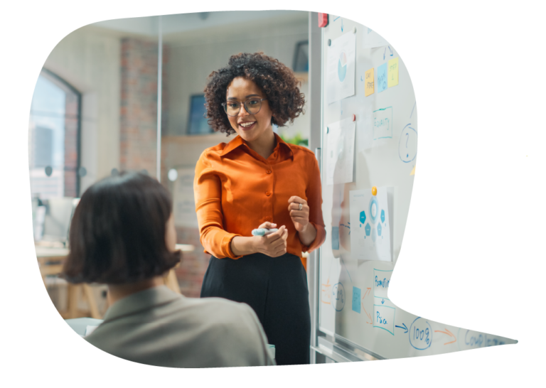 A business woman stands next to a full whiteboard speaking and gesturing to a seated person in an office.