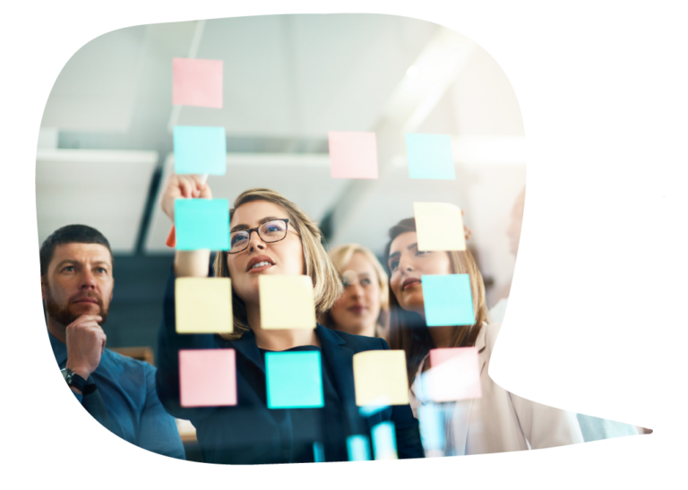 A woman stands facing a glass wall peppered with sticky notes as a group of business professionals stand behind her watching and looking thoughtful.