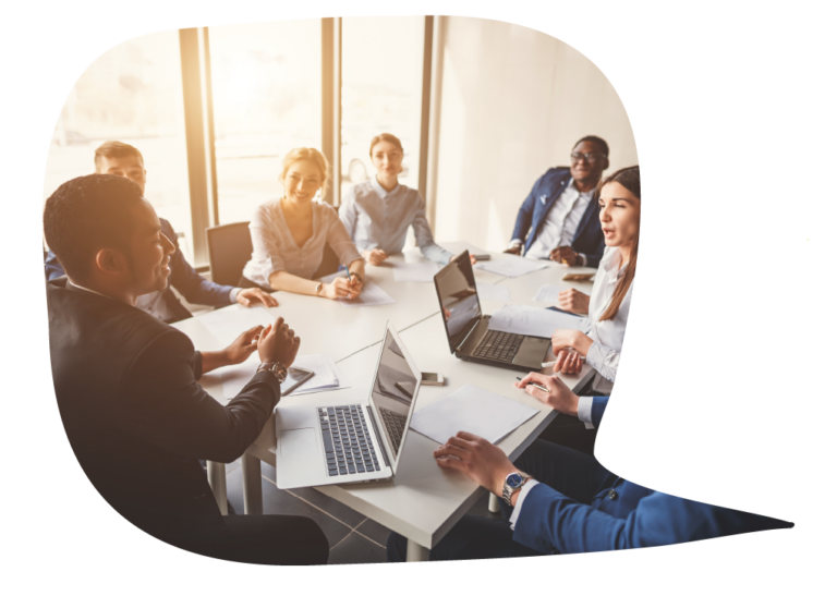 A person sits at the head of a table, leading the rest of the people around the table in listening and questions. There are several open laptops around the table and all are in business clothing.